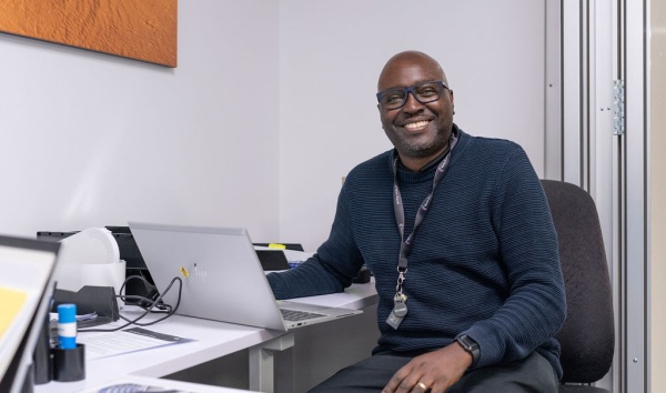 A man in a blue sweater sitting at a desk with a laptop and smiling into the camera