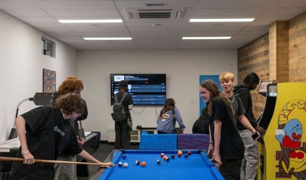 Some teenagers playing pool on a blue pool table while other teenagers hang out in the background