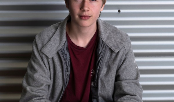 A boy in a red shirt and a grey jacket sitting down and looking into the camera