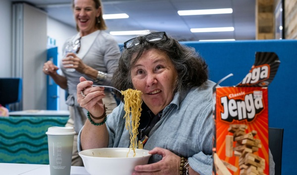 A woman making a funny face and eating noodles at a table while a woman behind her laughs