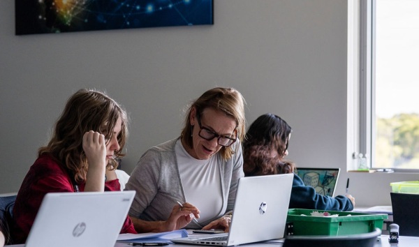 A woman sitting at a desk with a girl and explaining some schoolwork to her on a laptop