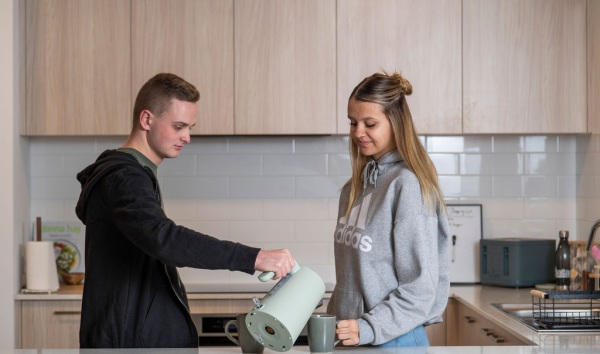 A teenage boy and teenage girl pouring tea for each other while standing at the kitchen counter