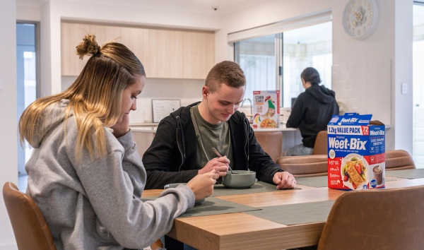 A teenage girl and teenage boy eating weet bix at the kitchen table while another girl eats corn flakes at the kitchen counter in the background