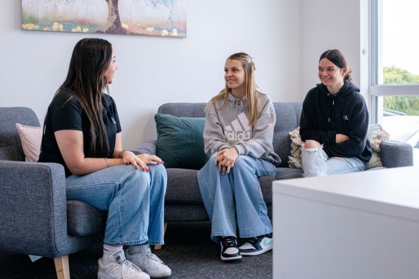 Three teenage girls sitting on some grey couches in a living room and chatting and smiling