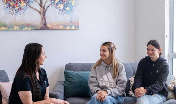 Three teenage girls sitting on couches in a living room and chatting with a painting of a tree in the background