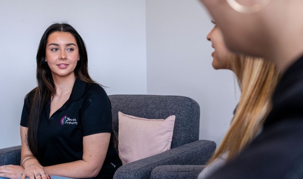A teenage girl in a youth futures polo listening to another teenage girl speak while sitting on a couch