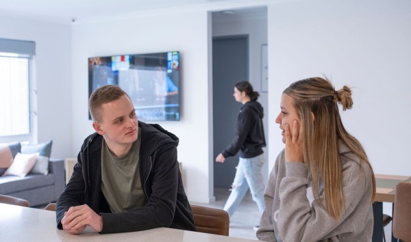 A teenage boy and a teenage girl sitting at a kitchen counter and talking while a girl in the background walks by in the background