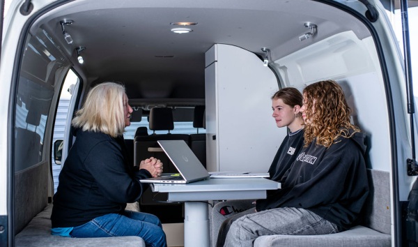 A woman sitting with two teenage girls at a desk inside a van with a laptop while they listen to her give some kind of explanation