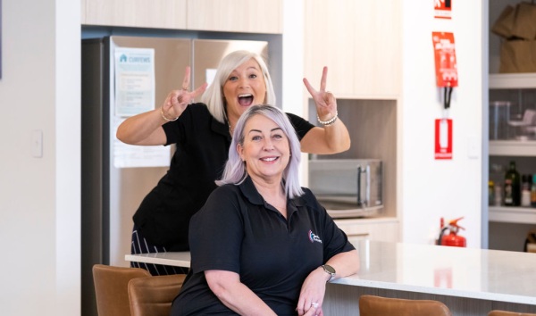 A woman sitting at a kitchen counter and smiling at the camera while the woman standing behind her makes an excited face and is doing the peace sign with both hands